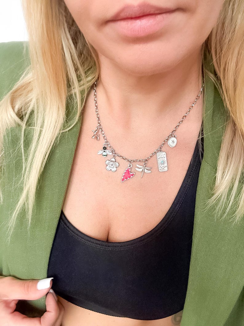 May include: A silver-toned charm necklace featuring multiple dangling pendants including a bee, a flower, a pink heart, a dragonfly, and a rectangular tag with celestial symbols. The chain is a standard link style with a secure clasp mechanism.