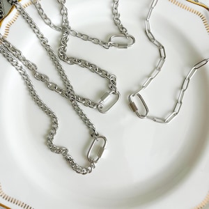 May include: Close-up of several silver-colored chain necklaces with carabiner clasps. The necklaces are displayed on a white plate with a gold trim. The chains have different link styles, offering a variety of options.