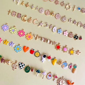 Könnte beinhalten: Eine Sammlung bunter Charms in verschiedenen Formen, darunter Blumen, Monde, Sterne und Sonnen. Die Charms sind gold-, silber- und emailliert, mit Designs wie Herzen, Äpfeln und Pilzen. Die Charms sind auf einer hellgelben Oberfläche angeordnet.