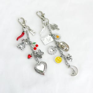 May include: Two silver-toned metal keychains featuring various charms. The left keychain includes a red chili, a letter B, a teddy bear, cherries, a heart, a butterfly, and a heart locket. The right keychain has a cactus, a postcard, a flower, a cowboy hat, a smiley face, and a crescent moon.