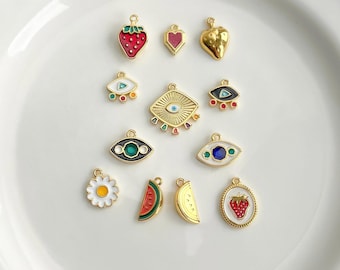 May include: A collection of small gold-toned metal charms featuring various enamel designs including a red strawberry, a pink heart, a gold strawberry, several evil eye motifs with colorful accents, a white daisy, and watermelon slices.