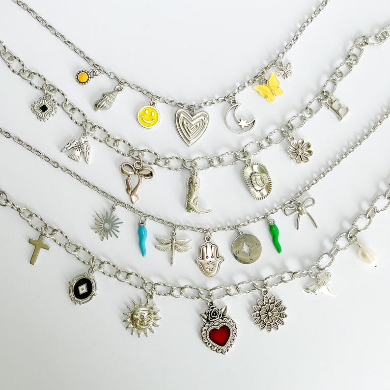 May include: Four silver-tone charm necklaces featuring various pendants including a yellow smiley face, a yellow butterfly, a heart, a crescent moon, a cowboy boot, a hat, a dragonfly, a hamsa hand, a cross, a sun, and a red heart.