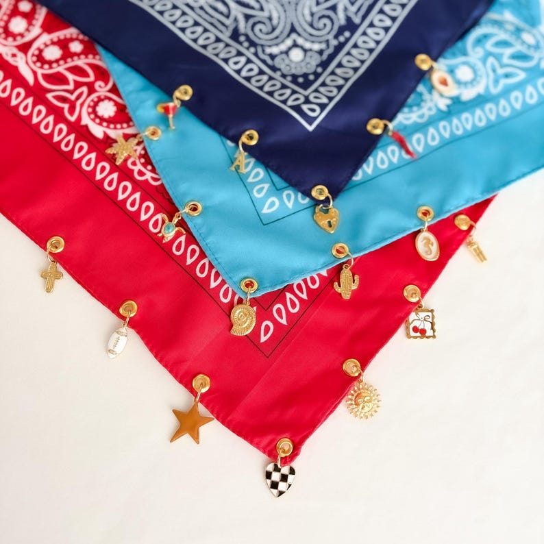 May include: Three layered bandanas in red, light blue, and navy blue, each featuring a white paisley pattern. The edges of the fabric are adorned with gold-toned metal grommets, from which various small charms hang, including a star, heart, and cactus.