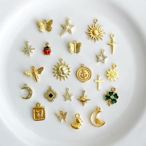 May include: A collection of gold-toned metal charms featuring various shapes including butterflies, stars, suns, a ladybug with red enamel, a bee, a crescent moon, a cross, an anchor, a duck, and a four-leaf clover with green enamel accents.