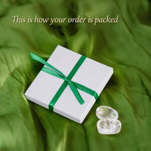 Op de afbeelding: Een vierkante witte geschenkdoos met een groen lint, vergezeld van twee heldere, gladde, afgeronde stenen. De tekst "This is how your order is packed" staat in een licht lettertype boven de doos gedrukt, tegen een achtergrond van groene stof.