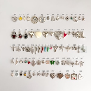 May include: A collection of metal charms arranged in rows and numbered from 81 to 155. The charms feature various shapes including hearts, stars, moons, animals, symbols, and food items, with some incorporating colored enamel accents.