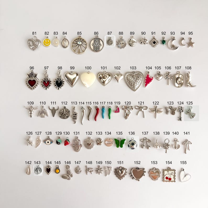 May include: A collection of metal charms arranged in rows and numbered from 81 to 155. The charms feature various shapes including hearts, stars, moons, animals, symbols, and food items, with some incorporating colored enamel accents.