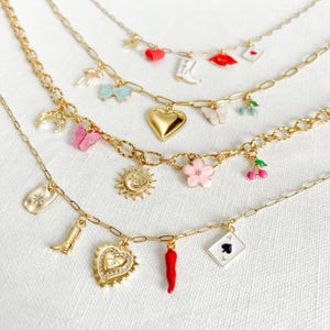 Op de afbeelding: Vier goudkleurige kettingen met diverse kleurrijke emaille en metalen bedels, waaronder een rood hart, een witte laars, rode lippen, een blauwe strik, een gouden hart, een roze vlinder, kersen, een zon, een roze bloem, een ster, een oog, een rode peper en een schoppen speelkaart.