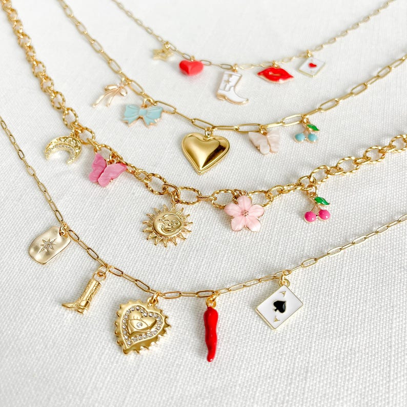 Op de afbeelding: Vier goudkleurige kettingen met diverse kleurrijke emaille en metalen bedels, waaronder een rood hart, een witte laars, rode lippen, een blauwe strik, een gouden hart, een roze vlinder, kersen, een zon, een roze bloem, een ster, een oog, een rode peper en een schoppen speelkaart.