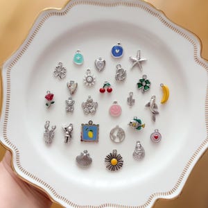 Könnte beinhalten: Eine Sammlung verschiedener Metallanhänger in unterschiedlichen Formen, darunter ein Stern, ein Herz, ein Seestern, eine Brezel, ein Kleeblatt, eine Rose, ein Bär, eine Blume, ein Smiley, ein Kaktus, ein Fisch, eine Banane, ein Hummer, ein Astronaut, eine Zitrone, ein Globus, eine Muschel und ein Gänseblümchen auf einem weißen Teller.
