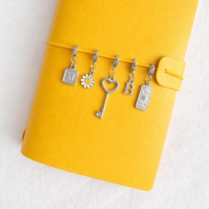 May include: A bright yellow notebook with a horizontal elastic band holding five silver-toned metal charms: a rectangular butterfly pendant, a daisy flower, a heart-shaped key, the letter B, and a rectangular sun-themed charm.