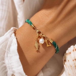 Op de afbeelding: Een goudkleurige bedelarmband met een groen en wit geweven band. De armband heeft een vissenbedel, een parel en de letter "L". De armband is vastgezet met een gouden sluiting.