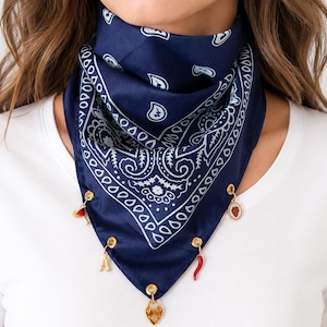 May include: A dark blue bandana featuring a white paisley pattern, adorned with five gold-toned metal charms attached to the bottom edge. The charms include a red cocktail glass, a strawberry, a gold letter A, a red chili pepper, and a gold heart.