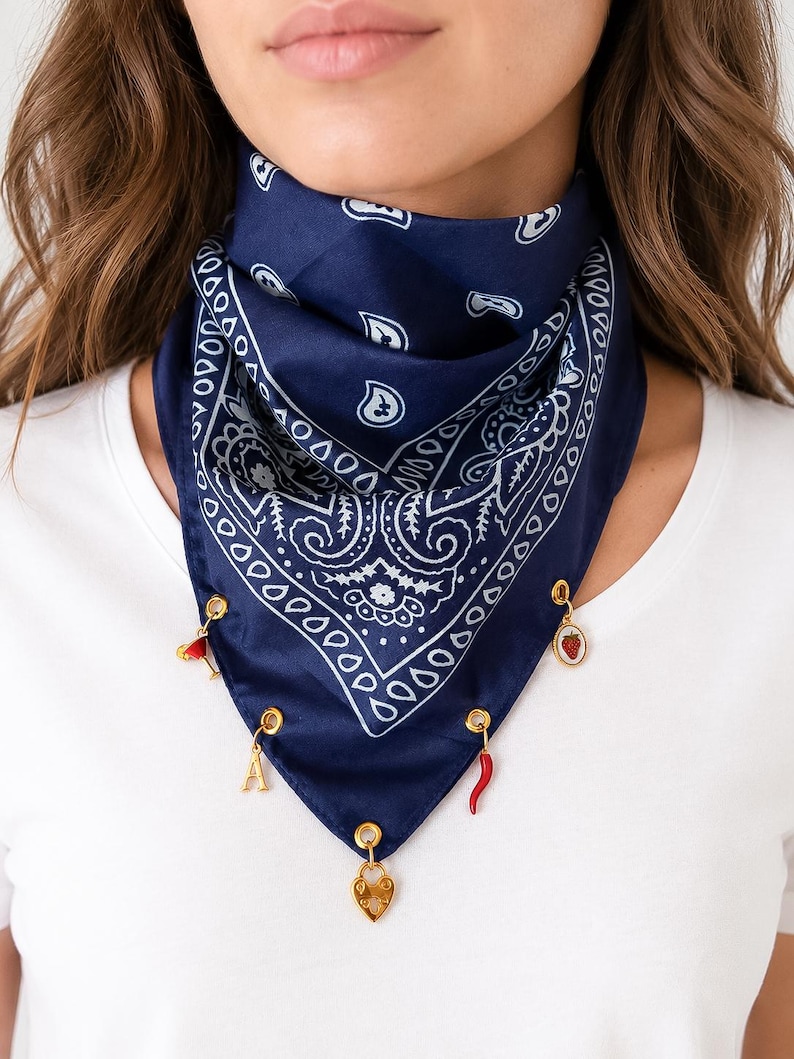 May include: A dark blue bandana featuring a white paisley pattern, adorned with five gold-toned metal charms attached to the bottom edge. The charms include a red cocktail glass, a strawberry, a gold letter A, a red chili pepper, and a gold heart.