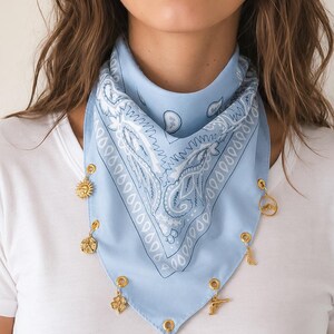 May include: A light blue bandana featuring a white paisley pattern and gold-toned metal grommets along the bottom edge. Several small gold-toned charms are attached to the grommets, including a sun, a star, a clover, a horseshoe, a boot, and a pistol.