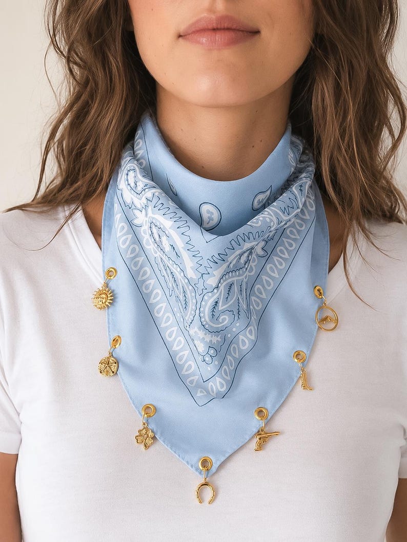 May include: A light blue bandana featuring a white paisley pattern and gold-toned metal grommets along the bottom edge. Several small gold-toned charms are attached to the grommets, including a sun, a star, a clover, a horseshoe, a boot, and a pistol.