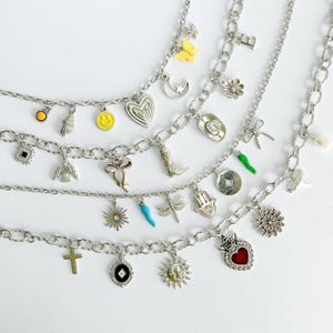 May include: Four silver-toned charm necklaces featuring various pendants including a yellow smiley face, a red heart, a cowboy boot, a dragonfly, a hamsa hand, a sun, a moon, a cross, a bow, a flower, and a letter E on a link chain.