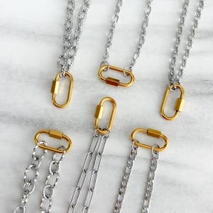 May include: A collection of silver-toned metal chains, each featuring a gold-toned oval carabiner clasp. The chains vary in link style, including twisted, oval, and flat link designs, all connected to the distinct gold-toned oval hardware.