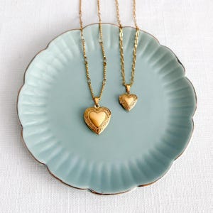 May include: Two gold-colored heart-shaped lockets on delicate chains. The lockets are displayed on a light blue, scalloped-edge plate. The larger locket has an ornate design around the heart. The chains are different styles.