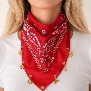 May include: A bright red bandana featuring a white paisley pattern. The bottom edge of the fabric is adorned with seven small gold-toned metal charms, including a sun, a mountain, a boot, a clover, a pistol, and a horseshoe, attached by rings.