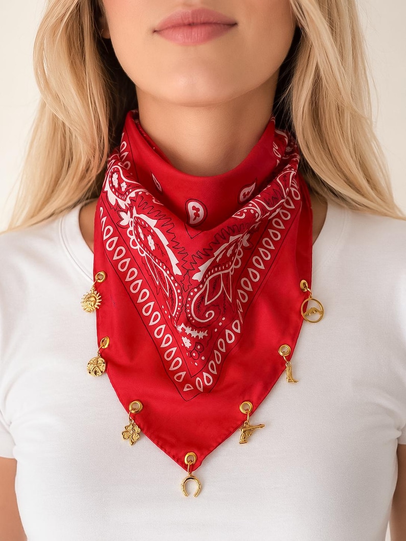 May include: A bright red bandana featuring a white paisley pattern. The bottom edge of the fabric is adorned with seven small gold-toned metal charms, including a sun, a mountain, a boot, a clover, a pistol, and a horseshoe, attached by rings.