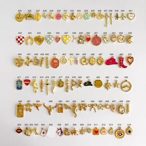 May include: A collection of gold-toned metal charms arranged in rows, each labeled with a number from 425 to 515. The charms feature various shapes including hearts, animals, symbols, food items, and geometric designs, some with colorful enamel accents.