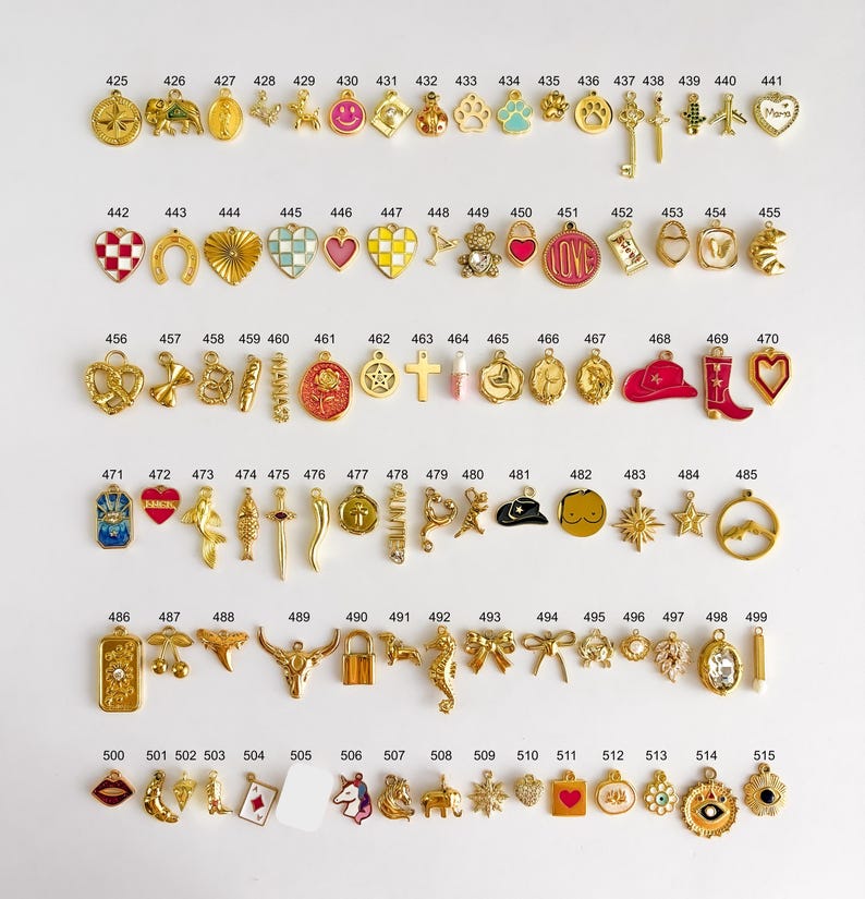 May include: A collection of gold-toned metal charms arranged in rows, each labeled with a number from 425 to 515. The charms feature various shapes including hearts, animals, symbols, food items, and geometric designs, some with colorful enamel accents.