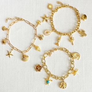 May include: Three gold charm bracelets featuring ocean-themed pendants like starfish, seashells, and suns. The bracelets have different chain designs and are presented on a white surface. The charms are detailed, offering a nautical aesthetic.