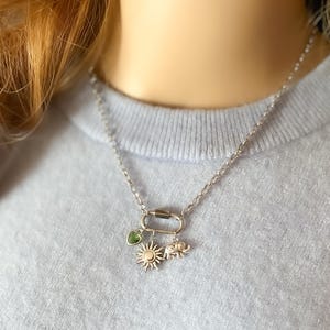 May include: A silver-toned chain necklace featuring a small oval carabiner clasp as a pendant. Attached to the carabiner are three charms: a small green heart-shaped stone, a detailed sunburst emblem, and a miniature elephant figure.