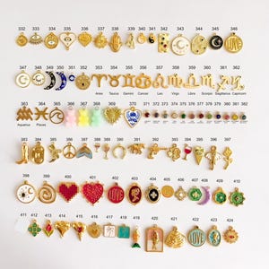 May include: A collection of gold-toned metal charms arranged in rows, numbered 332 through 424. The charms feature various shapes including celestial symbols, zodiac signs, hearts, flowers, animals, and colorful enamel accents on a white background.