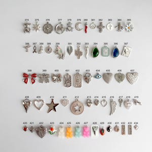 May include: A collection of metal charms arranged in rows, numbered 374 through 434. The charms feature various shapes including animals, flowers, hearts, stars, religious symbols, and geometric designs, with some incorporating colored enamel.