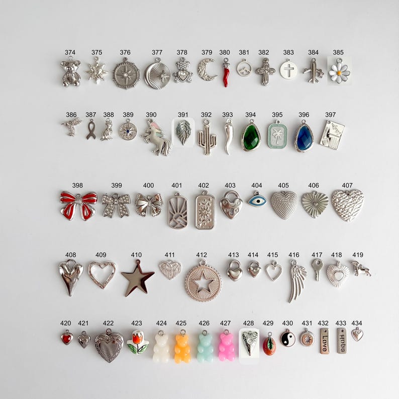 May include: A collection of metal charms arranged in rows, numbered 374 through 434. The charms feature various shapes including animals, flowers, hearts, stars, religious symbols, and geometric designs, with some incorporating colored enamel.
