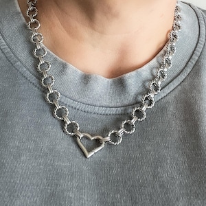May include: Silver-toned necklace featuring a heart pendant and a chain design. The necklace is composed of interconnected circular links and a heart-shaped centerpiece. The jewelry is worn against a gray shirt.