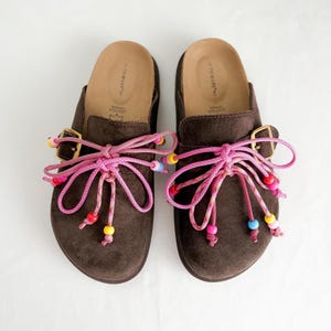 May include: A pair of dark brown suede slip-on shoes featuring gold-tone buckles and decorative pink cord laces tied in bows. The laces are adorned with small, multi-colored plastic beads in shades of red, yellow, blue, and pink at the ends.