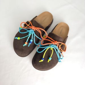 May include: A pair of dark brown suede slip-on clogs featuring adjustable side buckles and decorative lacing. The laces are bright orange, light blue, and neon yellow, accented with small, colorful plastic beads at the ends of the cords.