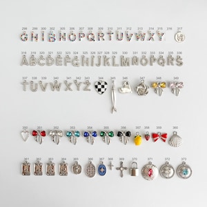 May include: A collection of metal jewelry charms arranged in rows, including multicolored rhinestone letters, silver-tone alphabet letters, a checkered heart, a fish, bows, fruits, crosses, and various circular pendants with numbers 298 to 373.