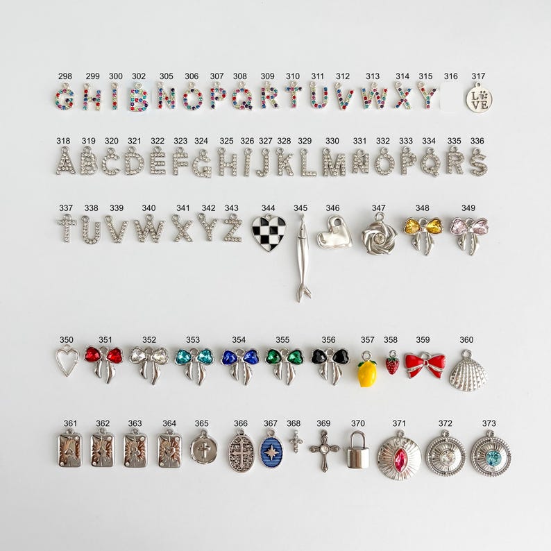 May include: A collection of metal jewelry charms arranged in rows, including multicolored rhinestone letters, silver-tone alphabet letters, a checkered heart, a fish, bows, fruits, crosses, and various circular pendants with numbers 298 to 373.