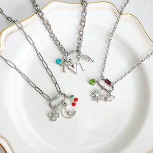 May include: Three silver-toned chain necklaces featuring carabiner-style clasps. Charms include a turquoise bead, letter T, heart, cross, wing, green bead, sun, elephant, four-leaf clover, crescent moon with star, and red cherries.