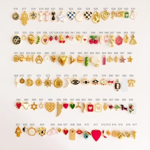 May include: A collection of small metal charms arranged in rows, each labeled with a number from 516 to 617. The charms feature various shapes including hearts, stars, animals, food items, symbols, and objects, finished in gold tones with enamel.