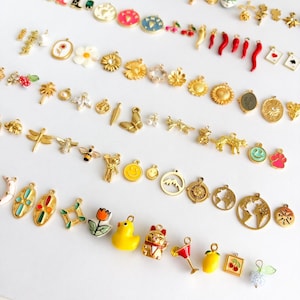 May include: A collection of assorted metal charms featuring various shapes including flowers, animals, food items, celestial symbols, and geometric patterns. The charms are finished in gold-tone metal, some with colorful enamel accents and small clear stones.