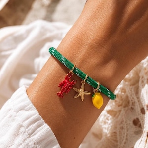 May include: A green cord bracelet featuring three dangling charms: a red lobster, a gold-toned starfish, and a yellow lemon. The cord has a white woven pattern throughout, and each charm is attached to the bracelet with a small gold-toned metal ring.