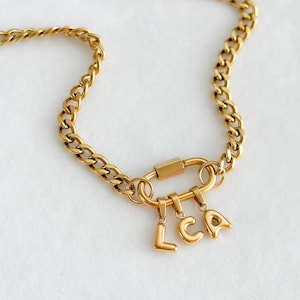 May include: Gold-coloured chain necklace featuring a large clasp and three hanging letter charms spelling "LCA". The chain has a curb link design. The necklace is displayed on a white surface.
