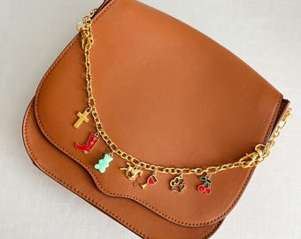 May include: A tan leather saddle bag featuring a gold-tone chain strap adorned with various charms, including a gold cross, a red boot, a light green gummy bear, a gold horse, a red wine glass, a gold paw print, and red cherries.