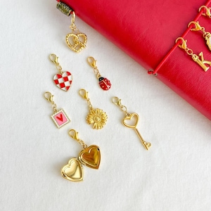 May include: A collection of gold-toned metal charms featuring a pretzel, a red and white checkered heart, a ladybug, a postage stamp with a red heart, a sunflower, a skeleton key, and an open heart-shaped locket, all with lobster claw clasps.