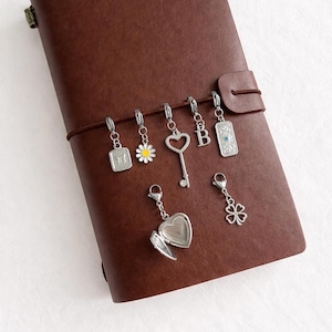May include: A dark brown leather journal with seven silver-toned metal charms attached to its elastic band. The charms include a rectangular butterfly tag, a daisy, a heart-shaped key, the letter B, a rectangular tag, a heart locket, and a clover.