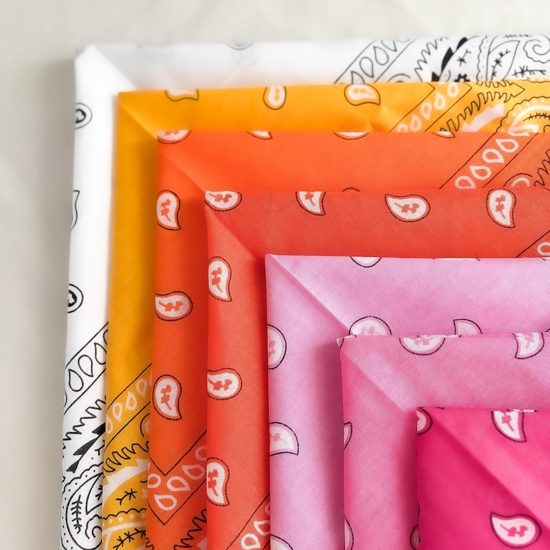 May include: A stack of folded square bandanas featuring a traditional paisley pattern. The collection includes a white bandana with black print, followed by yellow, orange, coral, light pink, and hot pink bandanas with white paisley motifs.