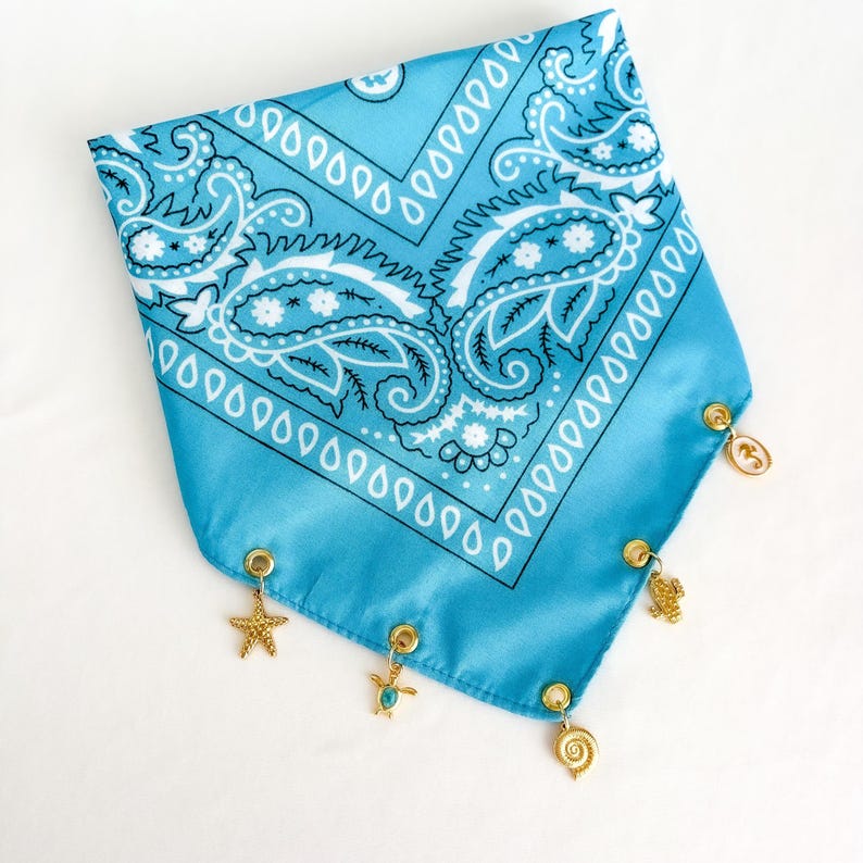May include: A light blue bandana featuring a white paisley pattern and a triangular shape. The bottom edge is adorned with five gold-toned metal charms, including a starfish, a sea turtle, and various shell-shaped designs attached by rings.