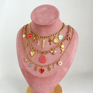 Op de afbeelding: Een gelaagde goudkleurige kettingenset met diverse bedels, waaronder een rood hart, een strik, de letter J, het woord MAMA, een kruis, een zon, een blauwe pootafdruk, een lieveheersbeestje, een bij, een roos, een roze ovaal hangertje en de letter B.