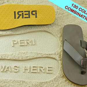 Peut inclure: Une paire de tongs marrons avec une semelle jaune. Les tongs sont sur une plage de sable avec les mots "PERI WAS HERE" imprimés dans le sable.