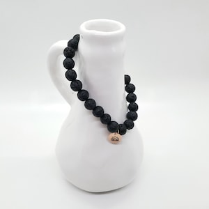 May include: Black lava rock beaded bracelet with a gold charm featuring a lotus flower design.